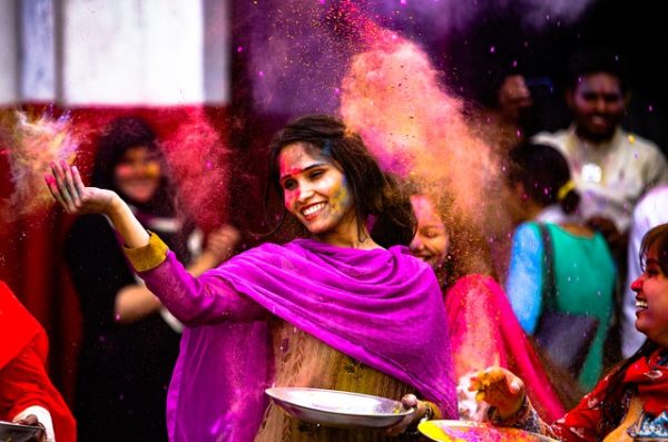 Woman celebrating what appears to be an Indian wedding or similar ceremony, wearing a bright scarf and people are throwing brightly colored smoke in the background.
