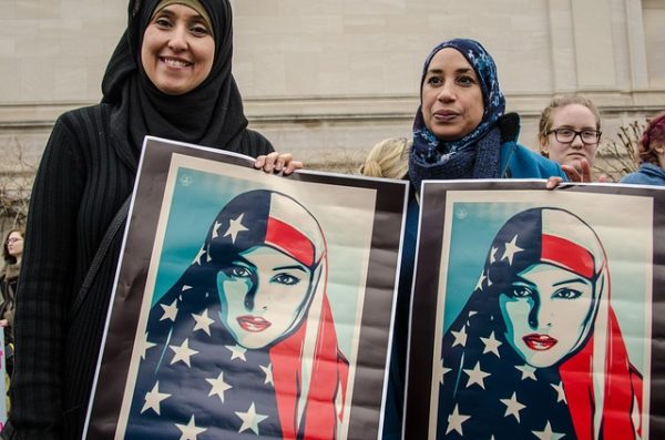 Muslim women holding paintings of muslim women clothed in American flag scarfs at what appears to be a protest.