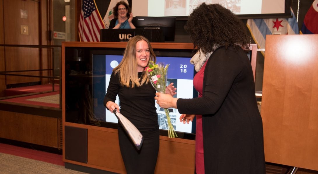 One person with long hair and a black sweater hands an award to a person in a black dress with blond hair.