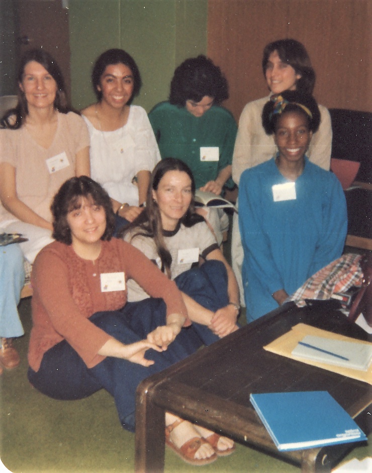 Seven members of the 1979 Women's Teaching Collective pose for a photograph while at the National Women's Studies Association. Four women are seated in a back row, all looking at the camera except the second woman from the right, who is looking down at a book in her lap. Three women are seated in a front row on the floor, which has olive green carpeting. A brown wooden coffee table stands in front of the front row, with a purse, folders, paper, and a pen on top of it..