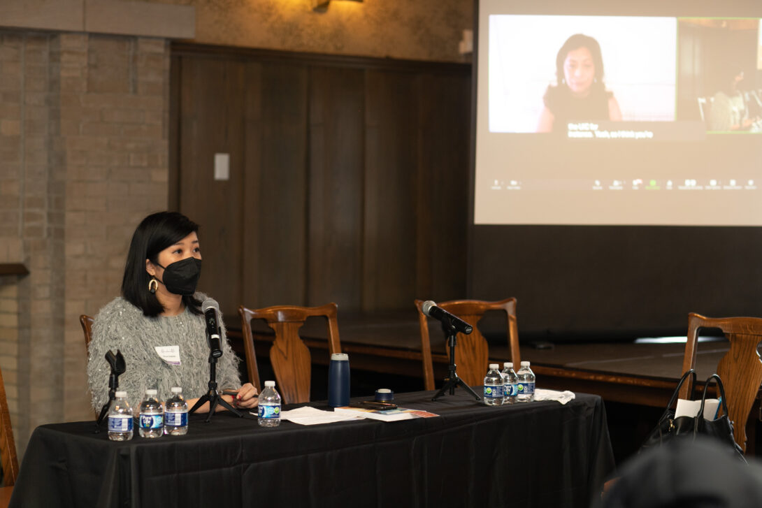 A panelist with shoulder-length black hear, wearing a gray sweater and black KN95 face mask, sits behind a table draped in a black tablecloth. Above and to the right of the table, a panelist participating via Zoom is projected on a screen. Small bottles of water, papers, and a microphone are on the table. Three empty wooden chars are behind and to the right side of the table.
