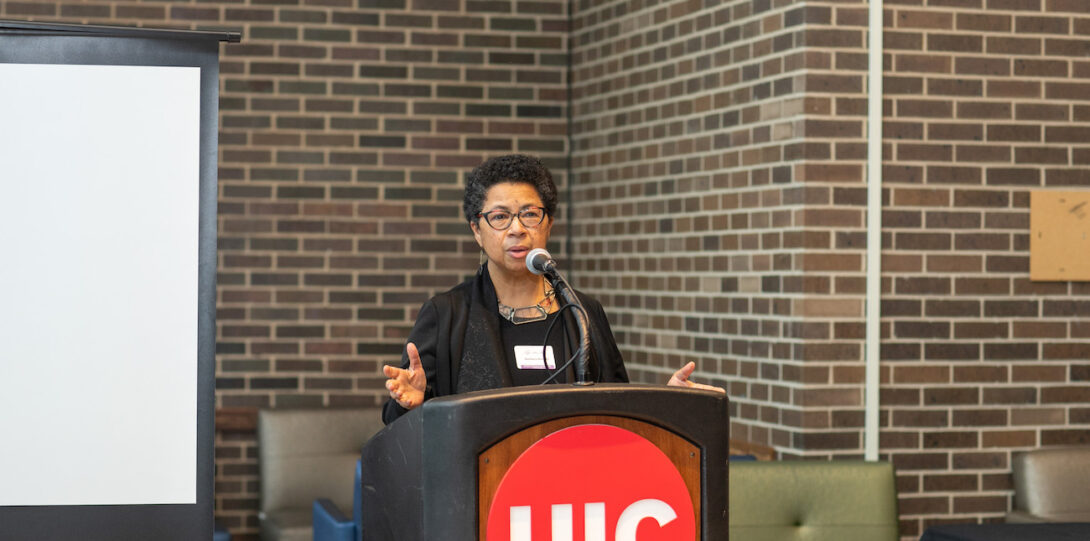 A woman with short black hair wearing glasses and a black dress stands behind a lectern with the UIC red circle logo with white font affixed to the front and speaks into a microphone atop the lectern.