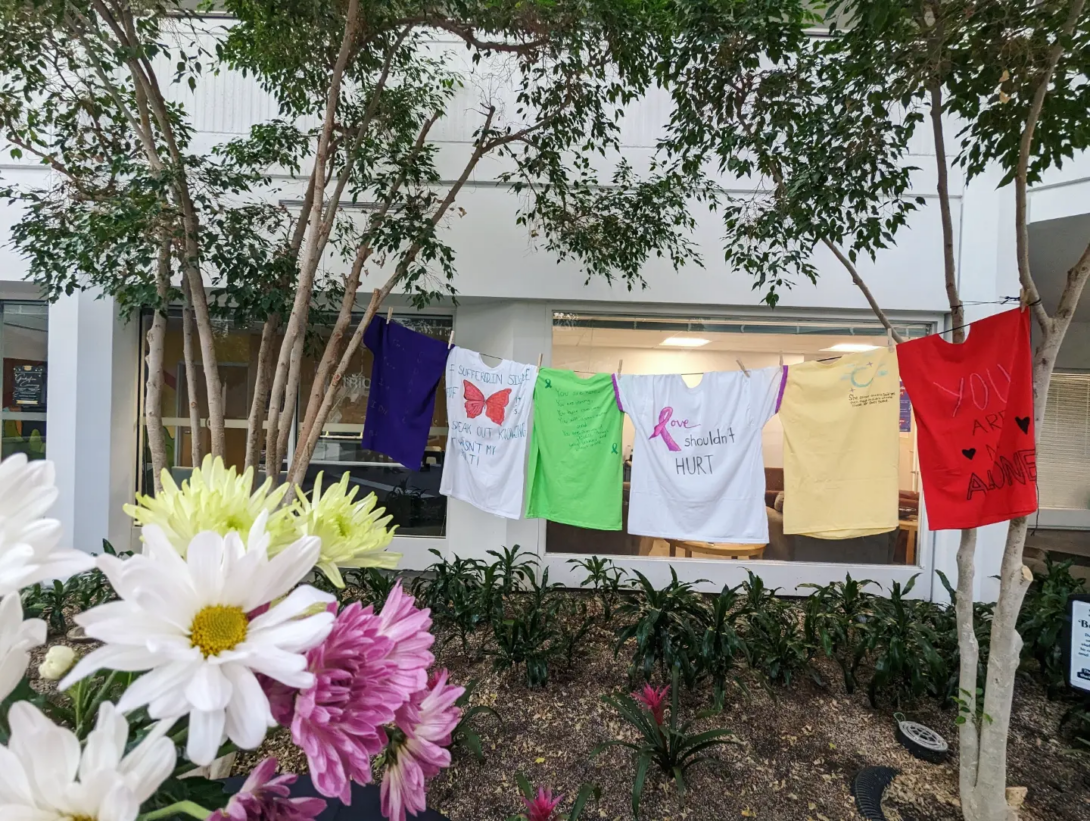 A row of t-shirts, titled the Clothesline Project, hand outside a white building in-between trees. There are white, yellow, and purple mums in front of the display.