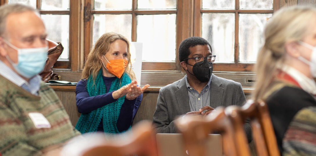 A woman with long blonde hair, wearing an orange face mask, dark blue sweater, and turquoise scarf, claps her hands while sitting. To her left sits a man with short black hair, wearing a black face mask, glasses, gray suit jacket, and a light gray button-down shirt.