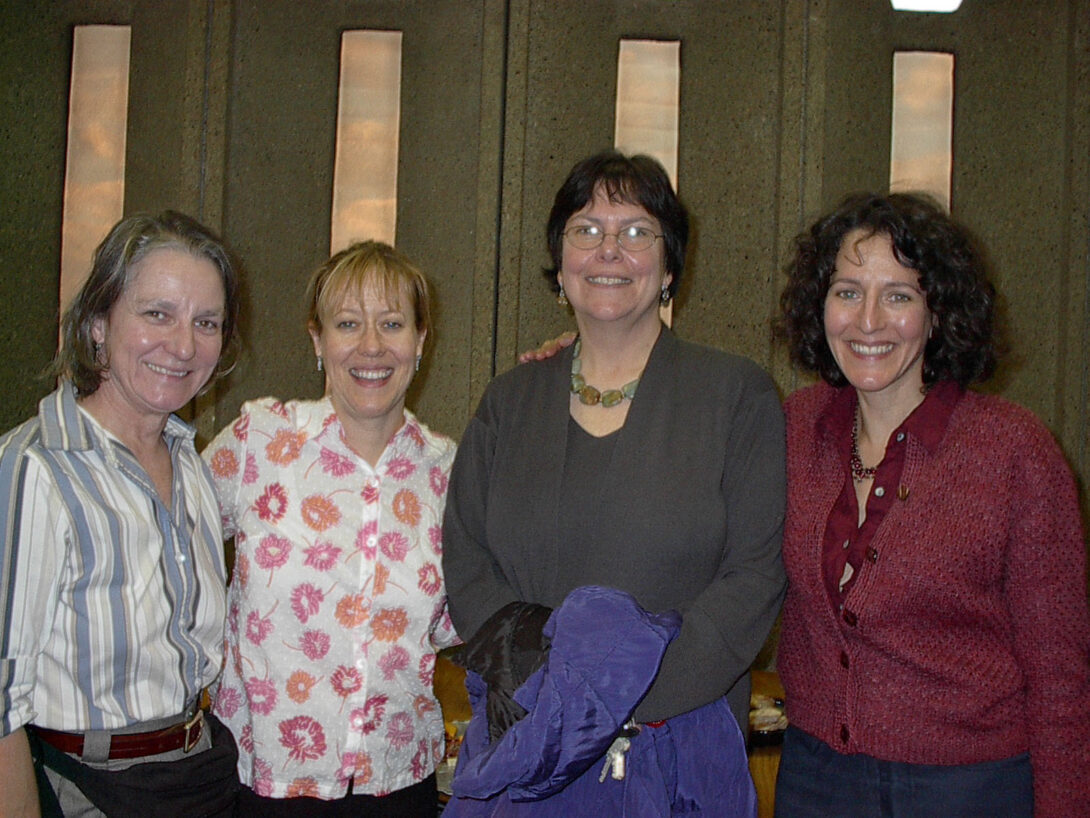 Four GWS faculty members stand in a row, smiling at the camera. They are in front of a wall in University Hall, with its iconic concrete slabs and long, narrow, vertical windows. Clouds and light blue sky are visible through the windows.