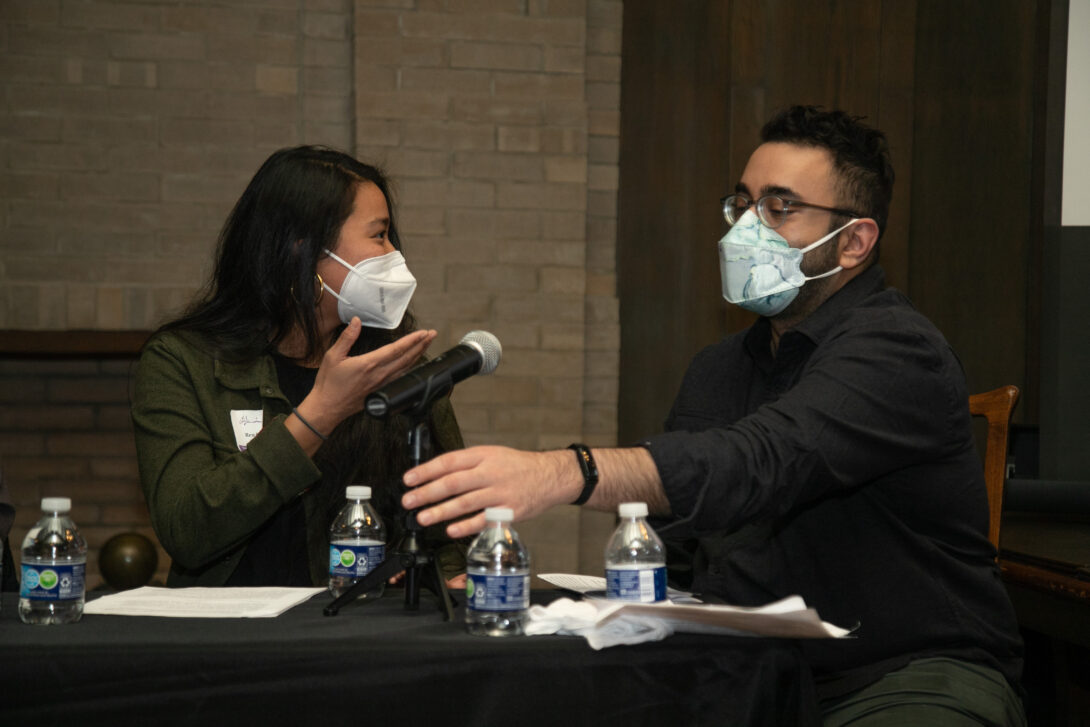 Two panelists sit behind a table draped in a black tablecloth. One panelist, a woman with long straight black hair wearing a face mask, motions to the panelist to her left, a man wearing glasses and a face mask and reaching for a microphone atop the table..