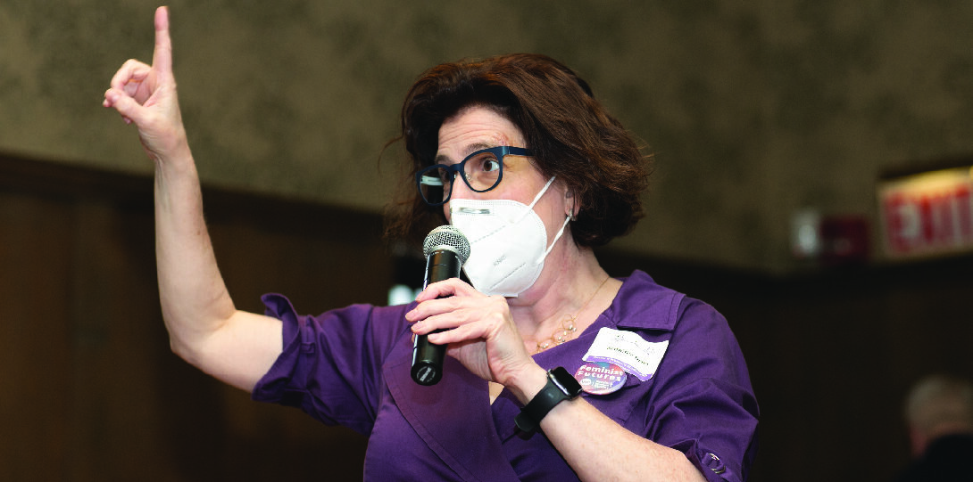 A woman with short brown hair wearing a purple dress, face mask, and glasses speaks into a microphone while raising her right arm and pointing her index finger toward the ceiling.