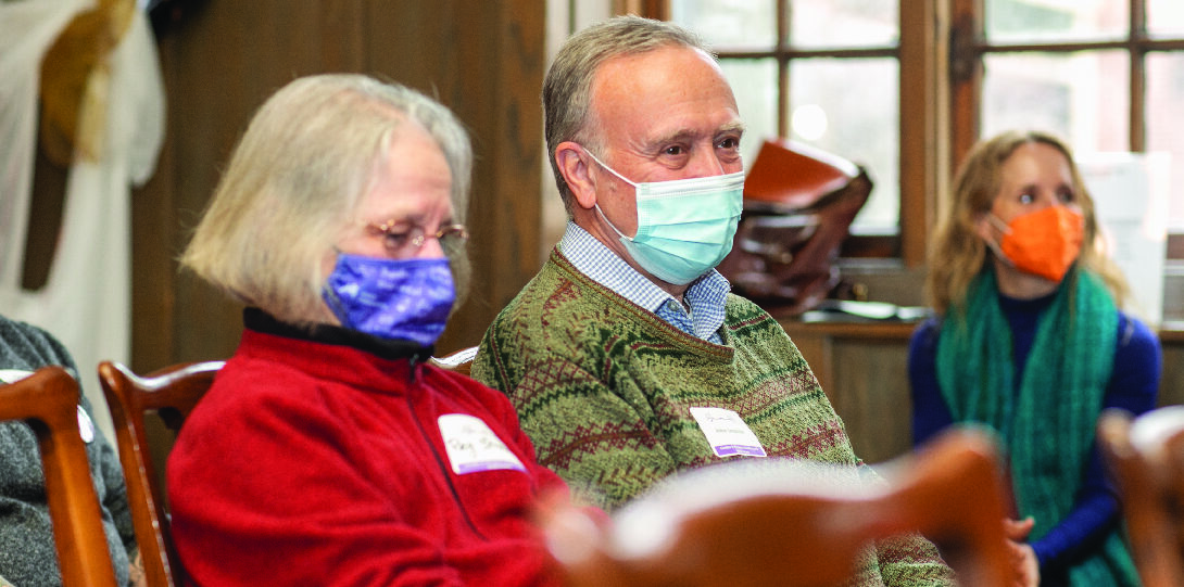 A woman with short gray hair wearing a purple face mask, glasses, and a red sweater sits next to a man with short gray hair, wearing a light blue face mask and a multi-colored sweater over a light blue collared shirt.
