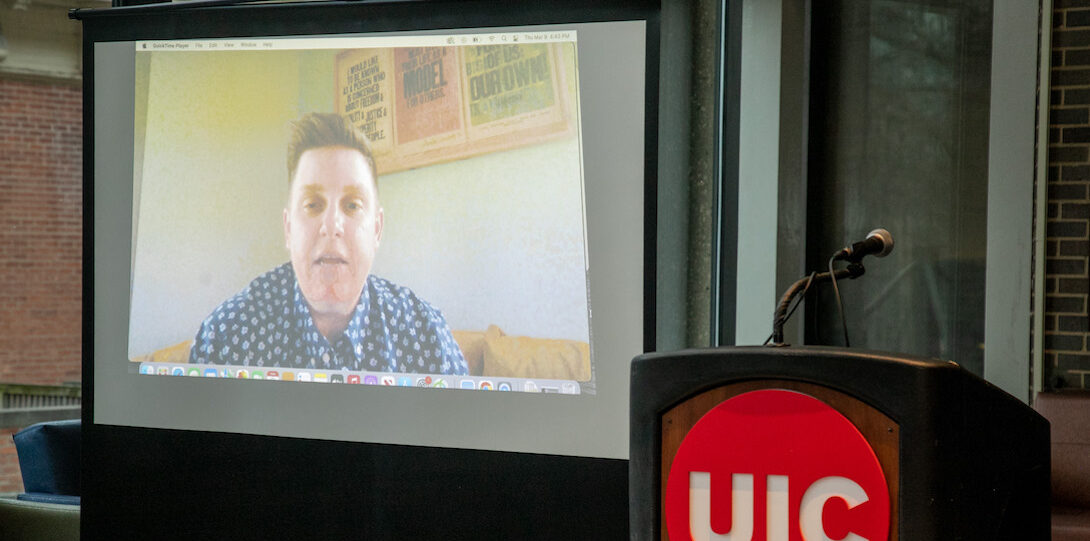 A video of a person with short light brown hair and a blue and white button-down collared shirt is projected onto a screen that stands to the right of a lectern with the UIC red circle logo with white font affixed to the front.