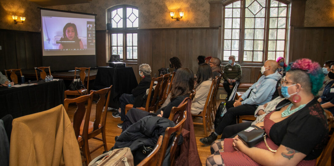 Attendees sit in wooden chairs arranged in rows and look toward a screen in the front of the room which displays a woman who is speaking to the audience via Zoom.