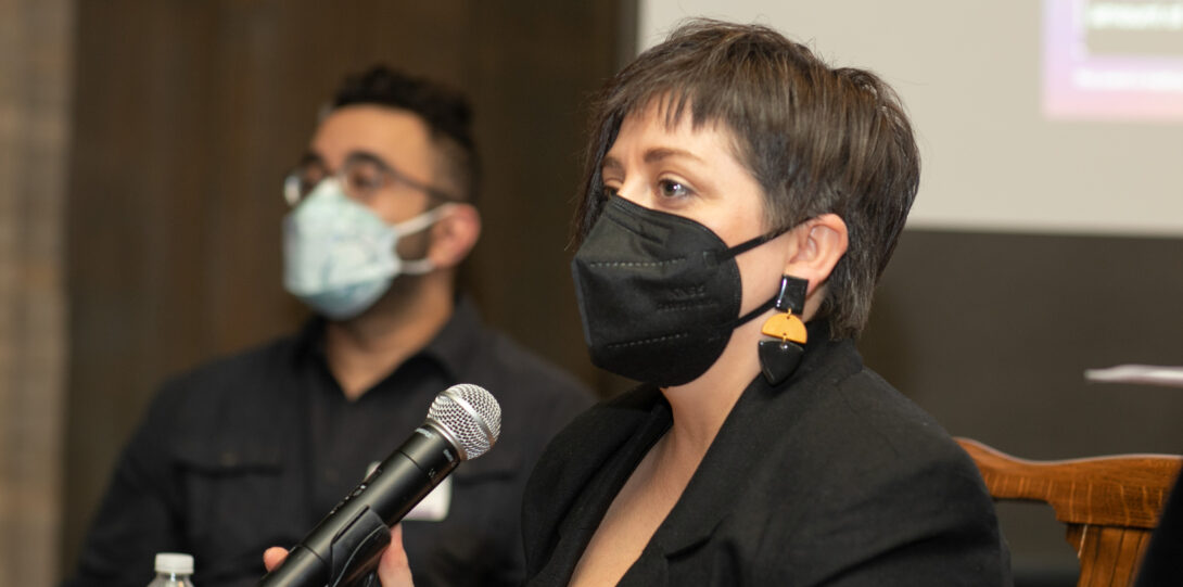 A woman with short brown hair wearing a black face mask, long black and beige earrings, and a black jacket over a beige shirt, speaks into a microphone.