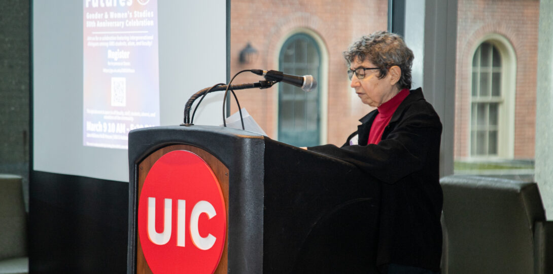 A woman with short gray hair wearing glasses and a black jacket over a red shirt stands behind a lectern with the UIC red circle logo with white font affixed to the front and speaks into a microphone atop the lectern.