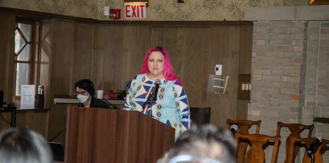 A woman with hot pink hair wearing a dress in shades of blue, white, black, and yellow and geometric shapes, stands behind a brown lectern and speaks into a microphone.