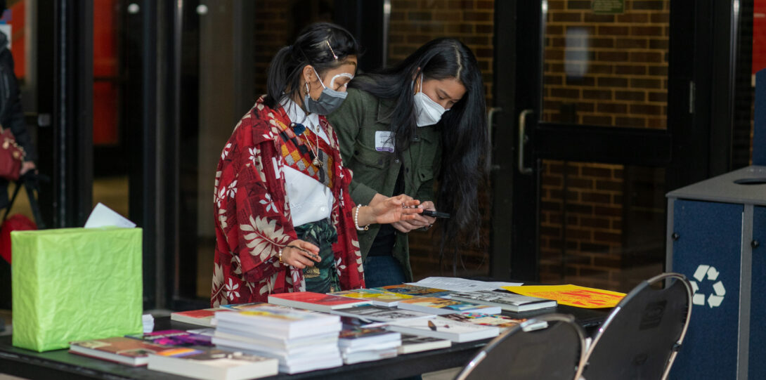 Two people stand side-by-side, looking down at a collection of books displayed on a table.