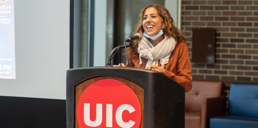 A woman with long reddish brown hair, wearing a thick beige scarf and an orange-ish brown jacket stands behind a lectern with the UIC red circle logo with white font affixed to the front and speaks into a microphone atop the lectern.