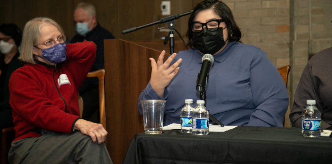 A woman with short gray hair, wearing a red sweater and gray pants, sits next to and looks at a woman with short black hair, who is wearing glasses, a black face mask, and a light blue shirt while speaking into a microphone.