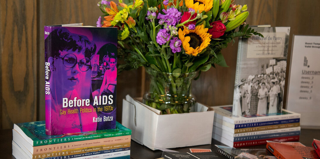 A colorful bouquet of flowers stands in a vase in the back center of a wood table. To the left is the book Before Aids, standing atop a stack of journals. To the right is the book Politics of the Pantry, standing atop a stack of journals. Fanned in front of the flowers are various books authored by GWS alum and faculty.