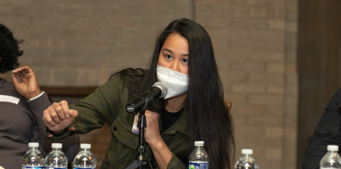 A person with long straight black hair wearing a white face mask and an olive green jacket over a black shirt speaks into a microphone.