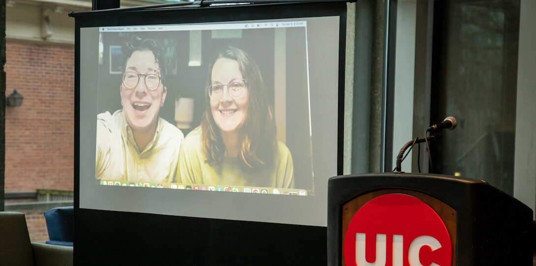 A video of two people sitting side-by-side is projected onto a screen that stands to the right of a lectern with the UIC red circle logo with white font affixed to the front.