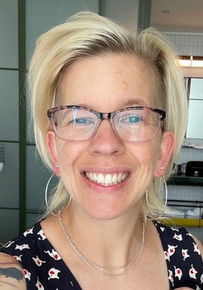 In this headshot, a woman with blond hair, wearing tortoise framed glasses, a blank tank top with a tiny white elephant print pattern, a silver necklace, and silver hoop earrings, looks at the camera and smiles.