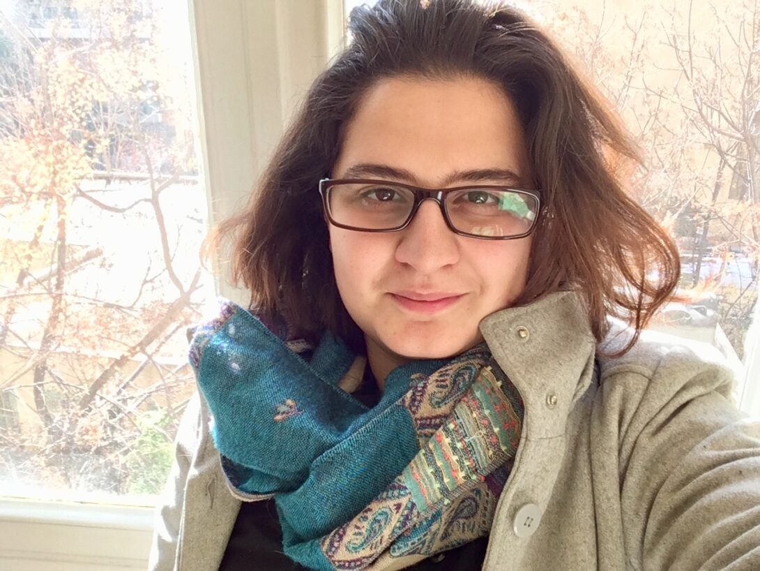 A person with glasses, brown hair, and blue scarf is in front of a window.