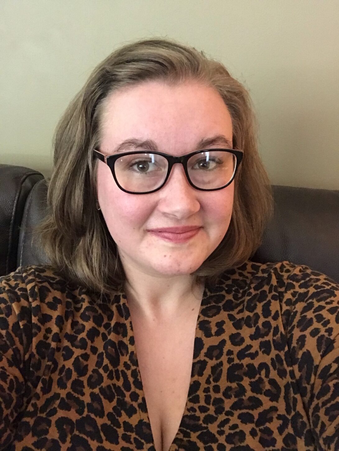 In this selfie, a person with glasses in a dark cheetah-print shirt sits on a couch in front of a wall.