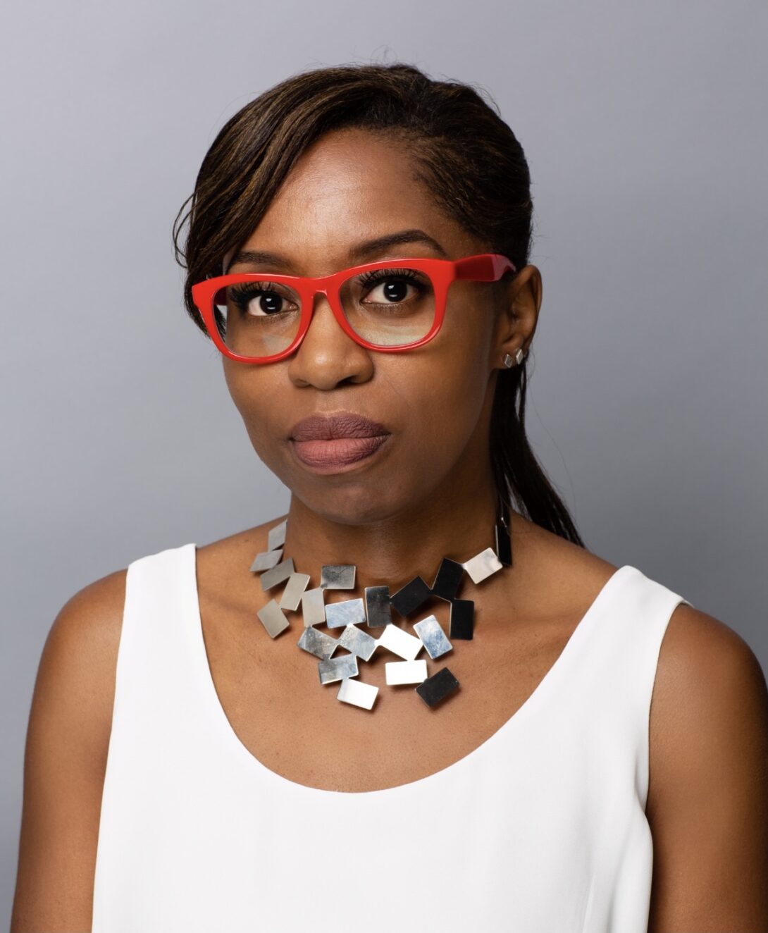 In this headshot, a person with a white tank top and red glasses looks at the camera.