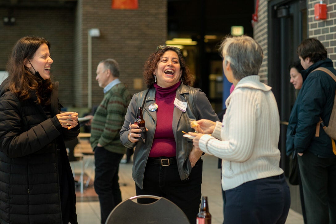 A group of people stand together with food and drinks in hand, laughing and having conversation.