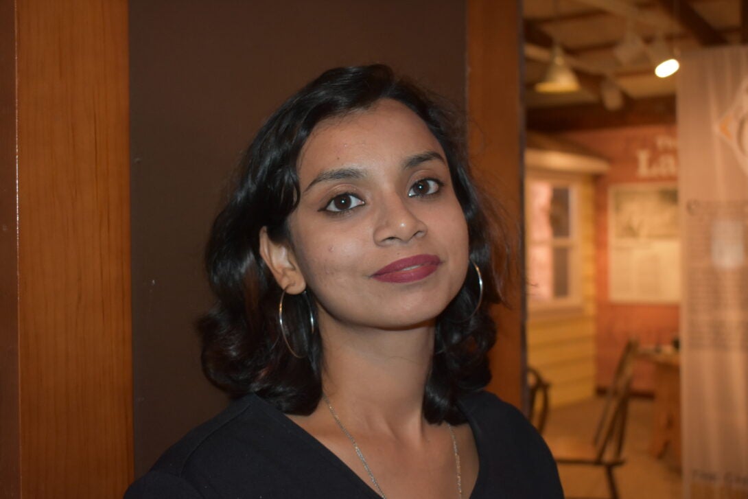 A person with brown skin and long brown hair is centered in this selfie, with a museum in the background.