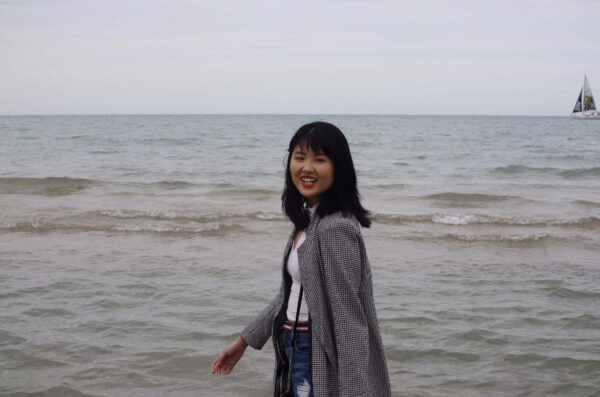 A person with black hair in a grey jacket stands in front of a body of water.