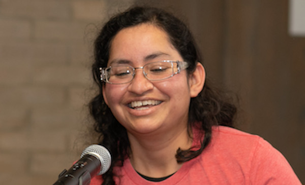 A person wearing glasses and a pink shirt smiles and speaks into a microphone.