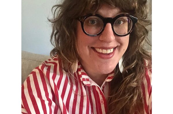 In this selfie, a person with long brown hair and glasses in a red and white striped shirt sits on a couch in front of a wall.