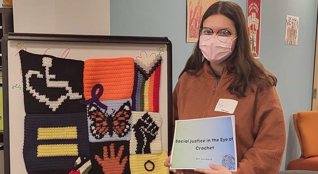 A woman with shoulder-length brown hair, wearing a light pink face mask, glasses, brown sweater, black jeans, and white gym shoes stands next to a crochet art piece she created. The rectangular crochet piece consists of smaller crochet pieces that each display a justice related symbol or message (ex. disability justice, reproductive justice, trans pride flag, migrant justice, black power fist, equality symbol). She is holding her artist's statement. On the cover page, it states 