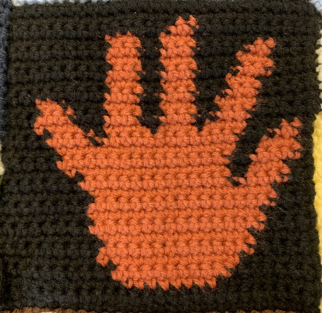 An orange crocheted hand appears on a black crocheted background