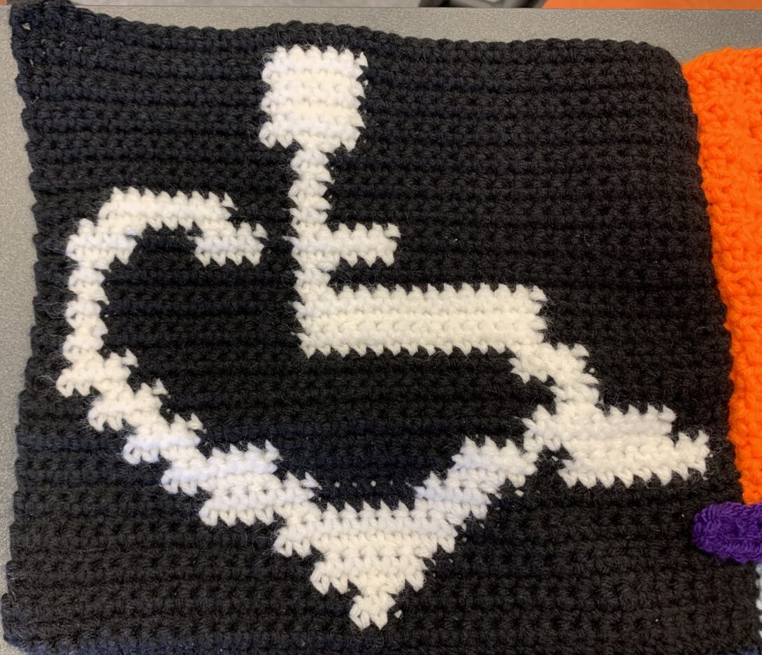 A black crocheted square piece. The wheelchair heart symbol is crocheted in white in the center of the square.