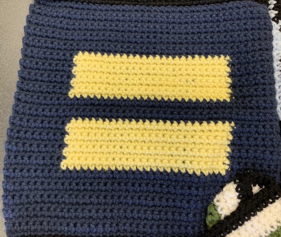 A yellow equality sign (two parallel horizontal yellow rectangles) is crocheted in the center of a navy blue crochet square