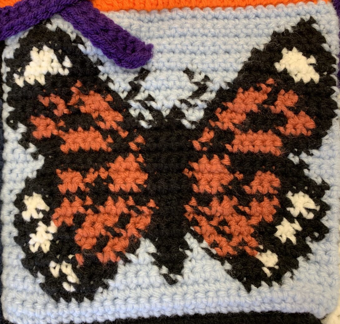 A monarch butterfly with black, orange, and white wings, is crocheted in the center of a light blue crochet square
