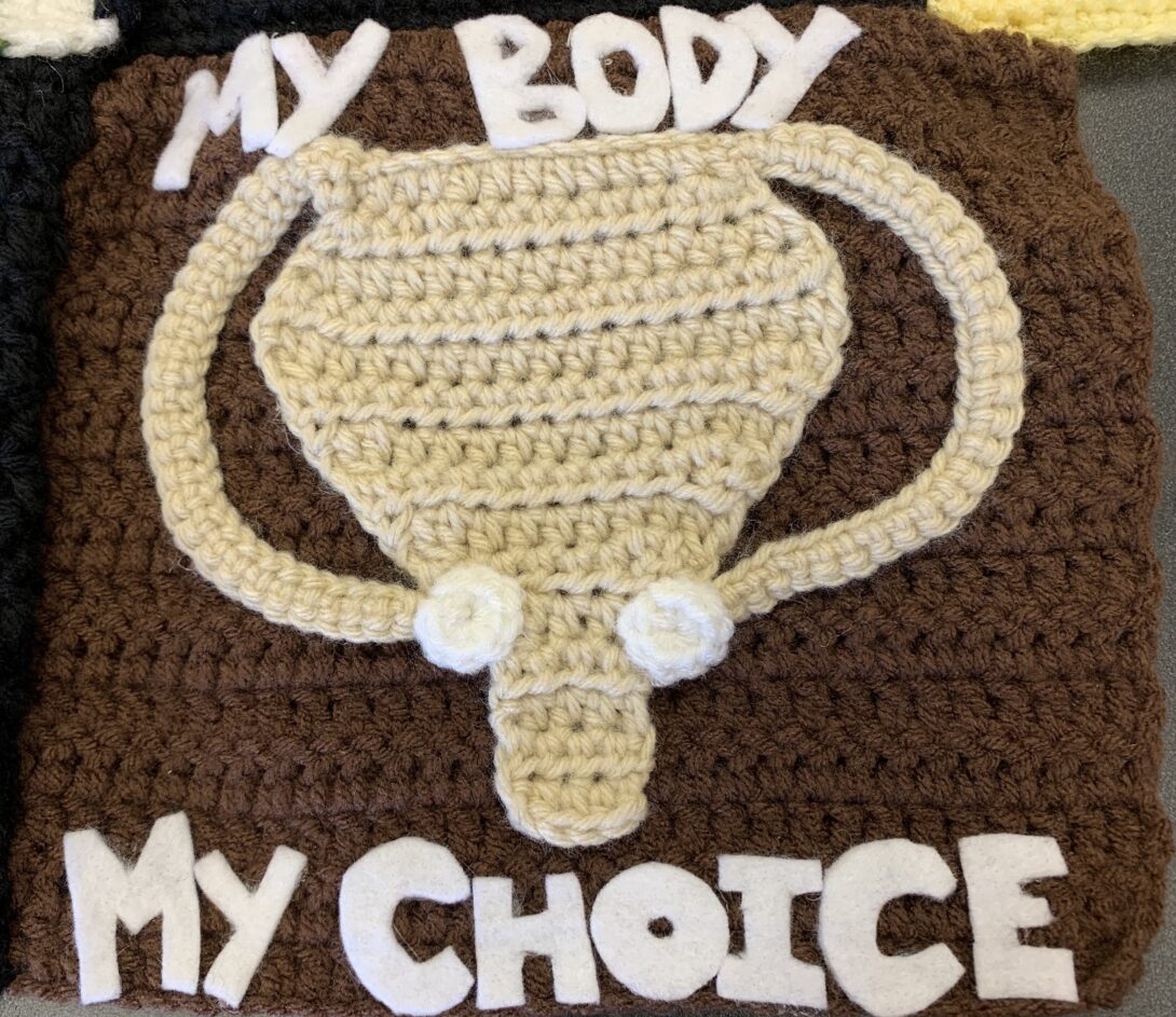 A brown crocheted square. In the center is beige crocheted uterus and fallopian tubes and white ovaries. At the top of the square, My Body appears in white felt letter. At the bottom of the square, My Choice appears in white felt letters.