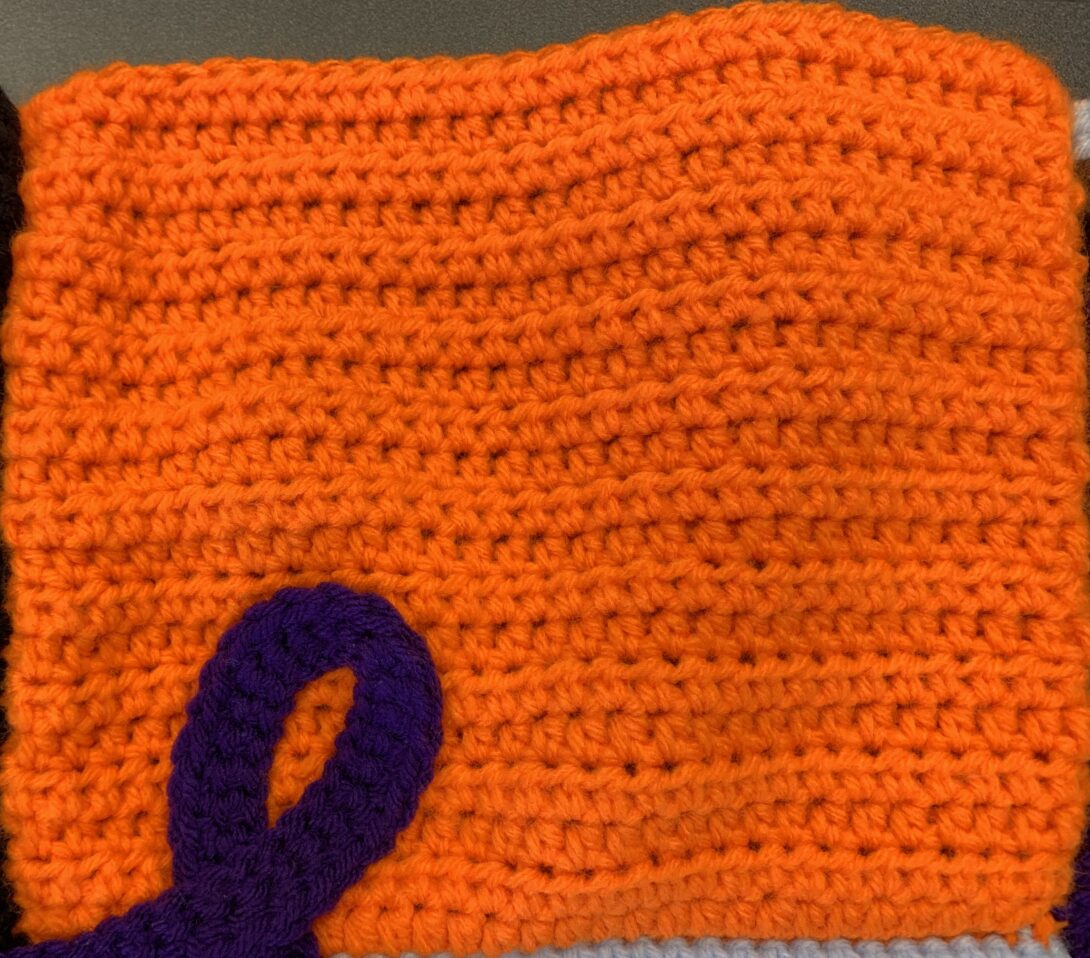 An orange crocheted square with the top of a purple crocheted ribbon in the bottom left corner.