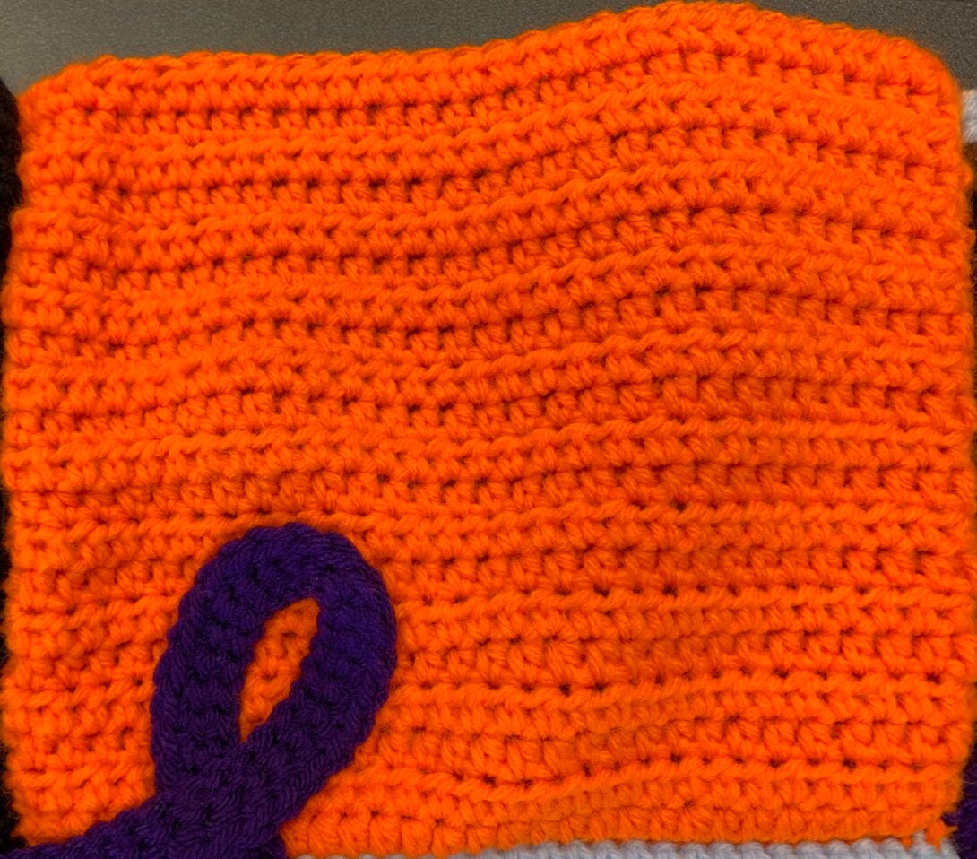 An orange crocheted square with the top of a purple crocheted ribbon in the bottom left corner.