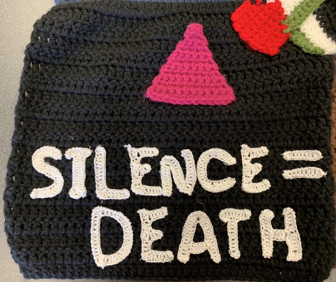 A pink crocheted triangle appears at the top center on a black crocheted background. Beneath the triangle, the phrase Silence = Death is crocheted in white.