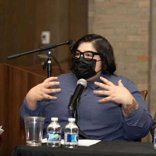 A person wearing a long sleeve, button-down light purple shirt, thick dark glasses, and a black face mask, with short straight black hair sits at a table speaking into a microphone