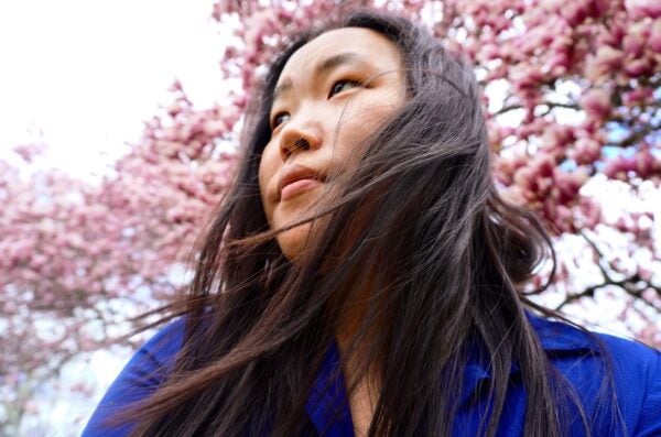 Photo of a person wearing a bright dark blue shirt with long, straight dark hair blowing in the wind. The photo looks up at the person, who is looking away from the camera toward their right. Behind and above the person is a tree with pink flower blossoms.
