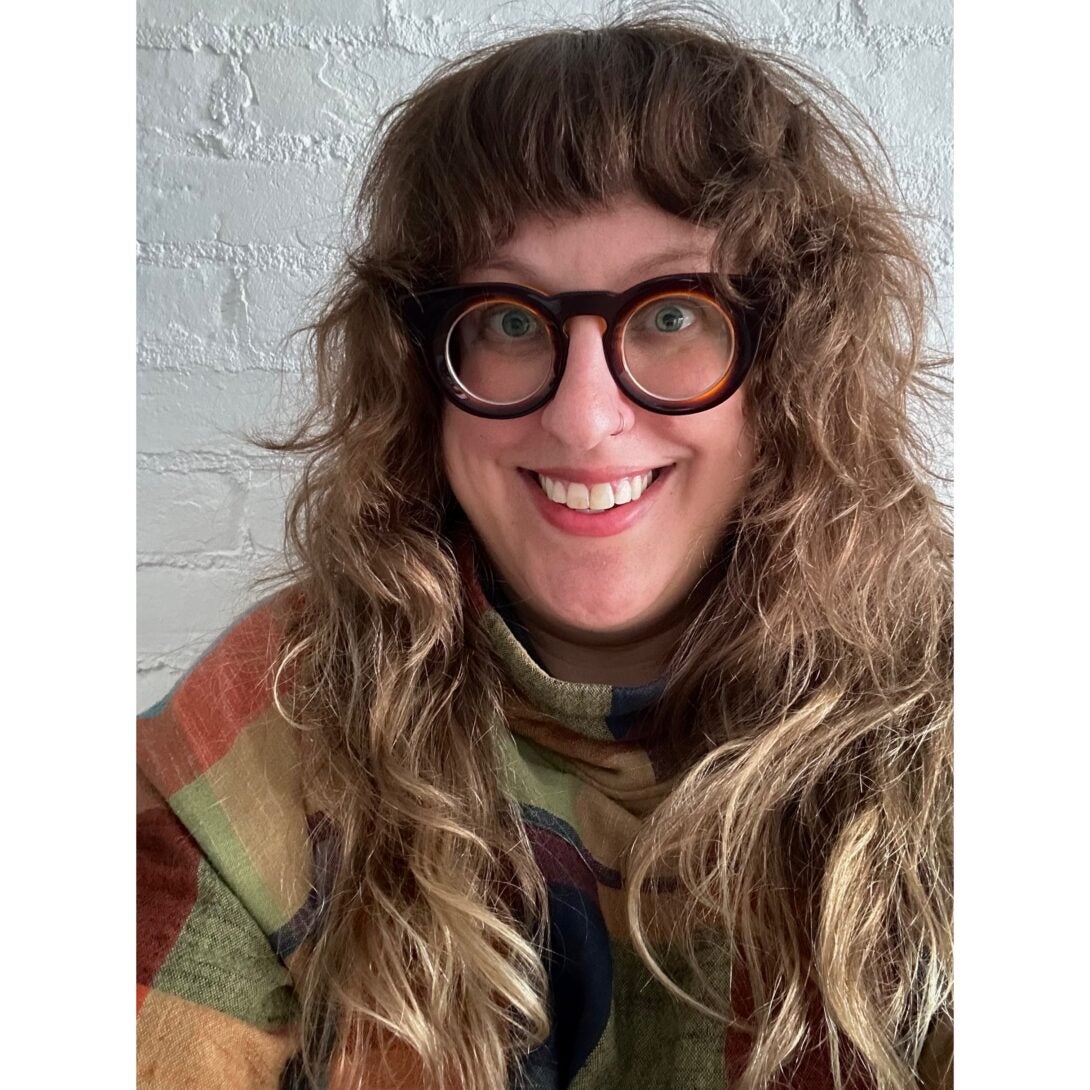 A person with long wavy blondish brown hair falling over their shoulders smiles broadly at the camera while wearing round brown glasses and a colorful checkered shirt.